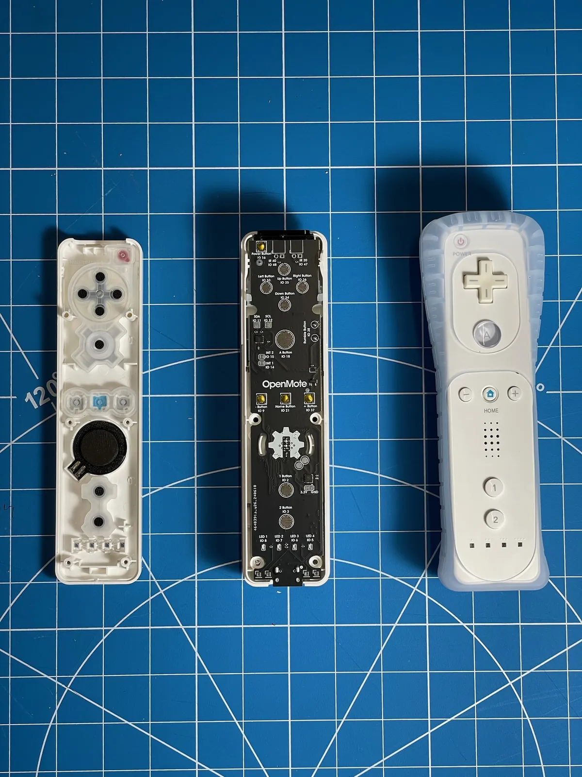 Three stages side by side on a cutting mat: an open Wii Remote shell with its rubber button pads, a black OpenMote board sitting in a white shell, and a finished remote